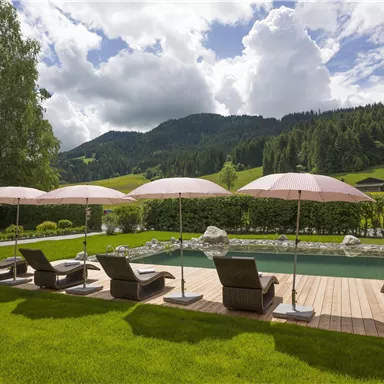 Ein schöner Garten mit Liegestühlen und Sonnenschirmen. Im Hintergrund sind Berge und wolkiger Himmel zu sehen.