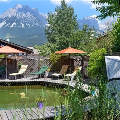 A cozy garden with a swimming pool and lounge chairs. In the background, impressive mountains and a blue sky can be seen.