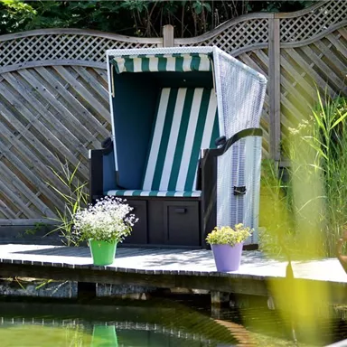 A cozy beach chair stands on a wooden terrace by the water. Around it are colorful flower pots with plants.