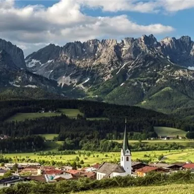 Eine malerische Landschaft mit hohen Bergen im Hintergrund und einem kleinen Dorf mit einer Kirche im Vordergrund. Der Himmel ist teilweise bewölkt und die Wiesen sind grün.