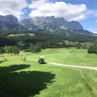 Eine malerische Golfanlage umgeben von Bergen. Der Himmel ist blau und es gibt viel grünes Gras.