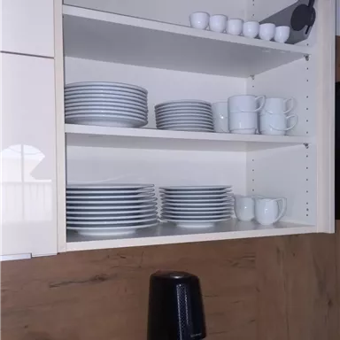 Ein geordneter Küchenschrank mit Tellern, Tassen und kleinen Schalen. Darunter befindet sich eine Kaffeemaschine und ein Informationsblatt.