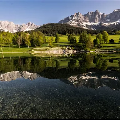 Ein ruhiger See spiegelt die majestätischen Berge und die grüne Landschaft wider. Der Himmel ist klar und strahlend blau.