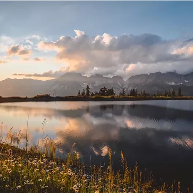 Eine ruhige Landschaft mit einem spiegelnden See und hohen Bergen im Hintergrund. Der Sonnenuntergang taucht die Szene in warmes Licht.