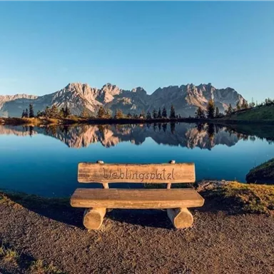 Eine gemütliche Bank steht am Ufer eines klaren Sees. Im Hintergrund sind majestätische Berge und ein blauer Himmel zu sehen.