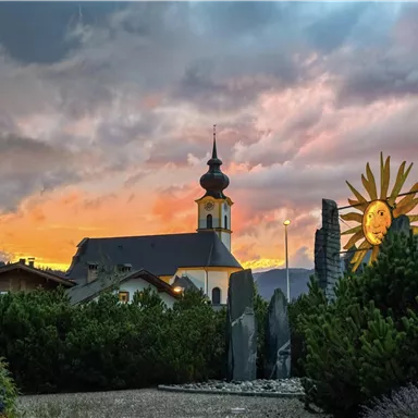 Eine schöne Landschaft mit einer Kirche vor einem farbenprächtigen Sonnenuntergang. Im Vordergrund sind grünes Pflanzenleben und eine Sonnenskulptur zu sehen.
