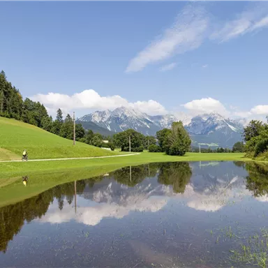 Eine ruhige Landschaft mit grünen Wiesen und Bergen im Hintergrund. Der Himmel ist blau und spiegelt sich im Wasser.