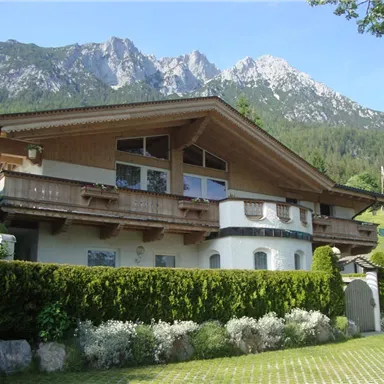 Ein malerisches Haus im alpenländischen Stil, umgeben von üppigem Grün. Im Hintergrund erhebt sich eine beeindruckende Berglandschaft.