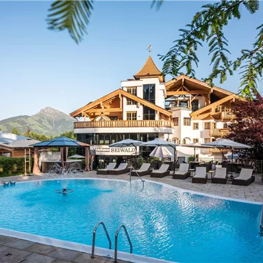 A pool in front of a charming hotel, surrounded by mountains and trees. The beautiful terrace invites you to relax.