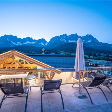 Eine atemberaubende Berglandschaft bei Dämmerung, mit modernen Liegen auf einer Terrasse. Im Hintergrund sind majestätische Berge und ein klarer Himmel zu sehen.