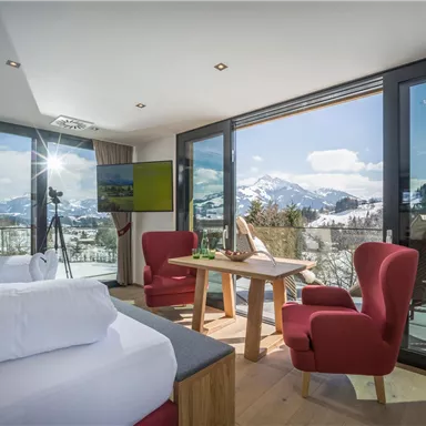 Ein modernes Zimmer mit großen Fenstern und Blick auf schneebedeckte Berge. Die Einrichtung umfasst zwei gemütliche rote Sessel und einen Tisch.