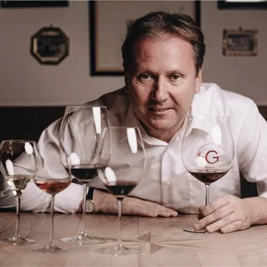 Ein Mann sitzt an einem Tisch mit mehreren Weingläsern. Er hält ein Glas Wein in der Hand und lächelt.