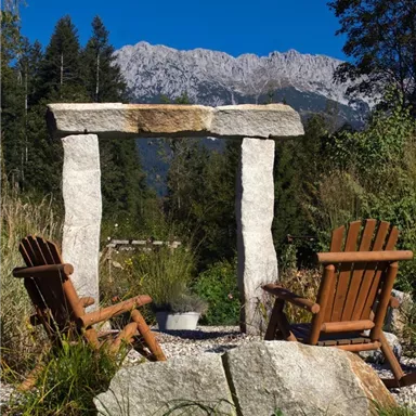 A tranquil garden with stone architecture and two wooden chairs. In the background, majestic mountains and a clear blue sky are visible.