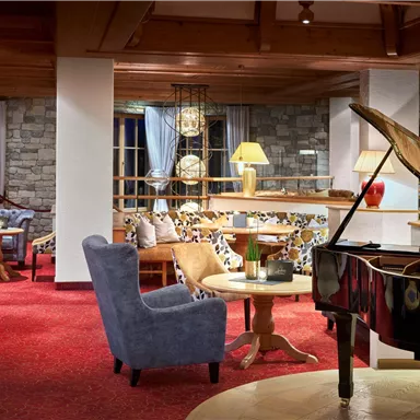 A stylish living room with a piano, cozy armchairs, and warm light. The walls are clad in stone and large windows let in plenty of daylight.