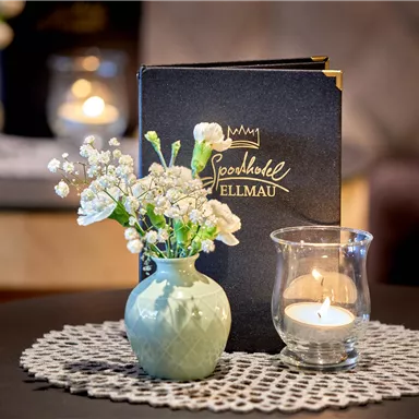 A small vase with white flowers stands on a table next to a candle and a menu. The table is equipped with a decorative placemat.