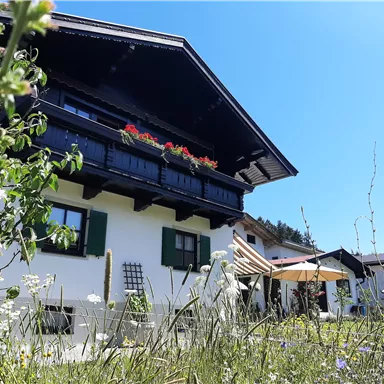 Ein traditionelles Haus mit einem Balkon voller Blumen und einem großen Garten. Die Sonne scheint an einem klaren blauen Himmel.
