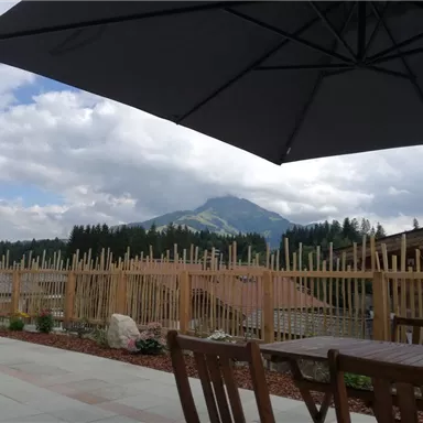 Ein schöner Ausblick auf einen Berg mit bewaldeten Hängen unter einem bewölkten Himmel. Im Vordergrund sind Tische und Stühle auf einer Terrasse zu sehen.