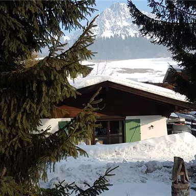 Eine gemütliche Berghütte im Schnee, umgeben von Fichten und mit einer majestätischen Bergkulisse im Hintergrund. Der Himmel ist klar und die Landschaft wirkt friedlich.