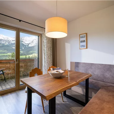 Ein gemütlicher Essbereich mit einem Holztisch und einer Bank. Im Hintergrund sind große Fenster, die eine schöne Aussicht auf die Berge bieten.