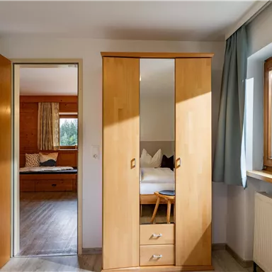 Ein helles Zimmer mit einem Holzschrank und Fenstern, die Tageslicht hereinlassen. Im Hintergrund ist ein weiterer Raum mit einem Bett und einer Aussicht auf die Natur zu erkennen.