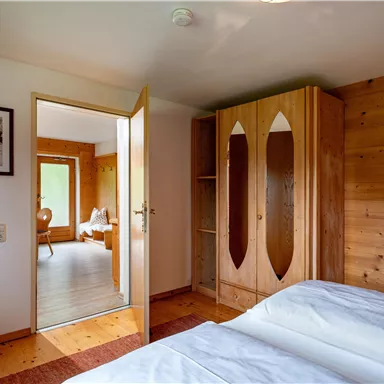 Ein gemütliches Schlafzimmer mit zwei Betten und einem Holzschrank. Im Hintergrund ist ein offenes Zimmer sichtbar.