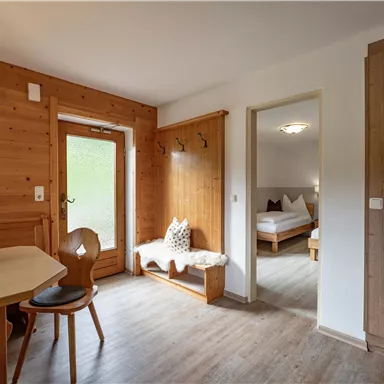 Ein gemütlicher Wohnbereich mit Holzwänden und einem Tisch. Im Hintergrund ist ein Schlafzimmer mit zwei Betten sichtbar.
