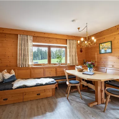 Ein gemütlicher Raum mit Holzvertäfelung und großen Fenstern. Am Tisch steht ein Blumenarrangement und eine Sitzecke ist vorhanden.