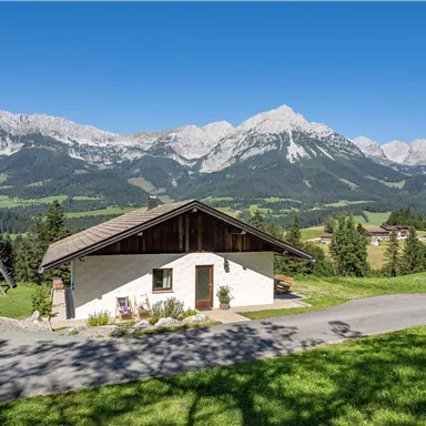 Ein gemütliches Haus in einer malerischen Berglandschaft. Im Hintergrund erheben sich majestätische Berge unter einem blauen Himmel.