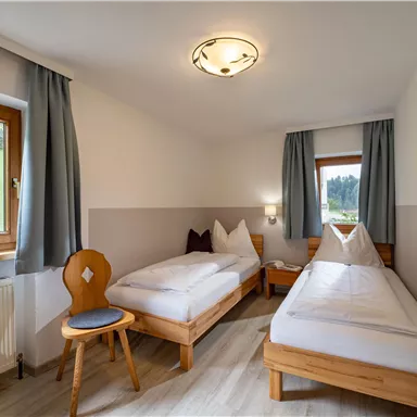 Ein einfaches Zimmer mit zwei Einzelbetten, einem Stuhl und einem Fenster mit Blick ins Freie. Die Wände sind hell gestrichen und die Fenster haben Vorhänge.