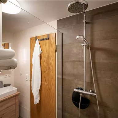 Ein modernes Badezimmer mit einer Regendusche und einer Holzdoor. Das Waschbecken ist einfach und elegant gestaltet, neben einem Handtuch und einem Bademantel.