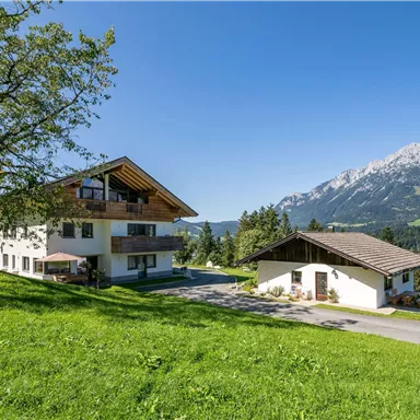 Eine malerische Landschaft mit einem großen Haus und einem kleineren Gebäude. Im Hintergrund sind majestätische Berge und ein klarer blauer Himmel zu sehen.