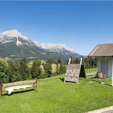 Eine idyllische Landschaft mit einer Wiese, einem Klettergerüst und einem Sitzplatz. Im Hintergrund erheben sich majestätische Berge unter einem klaren blauen Himmel.