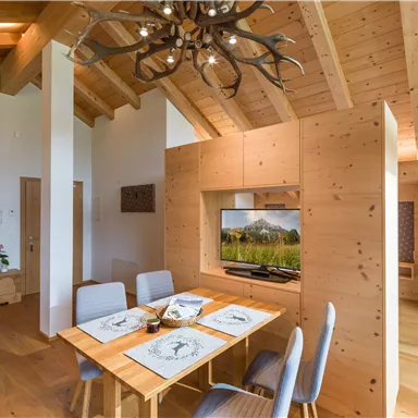 Ein helles und modernes Esszimmer mit Holzdekor und einem großen Tisch. Im Hintergrund ist ein Fernseher mit einem Bergpanorama zu sehen.