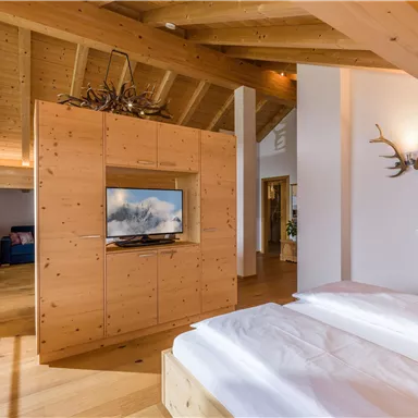 Ein modernes Schlafzimmer mit Holzdecken und einem gemütlichen Holzschrank. Ein Fernseher ist in die Möbel eingebaut, und es gibt große Fenster, die viel Licht hereinlassen.