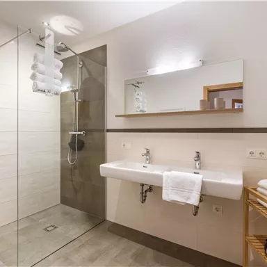 Ein modernes Badezimmer mit einer großzügigen Dusche und einem eleganten Waschbecken. Handtücher sind ordentlich auf einer Holzablage gestapelt.