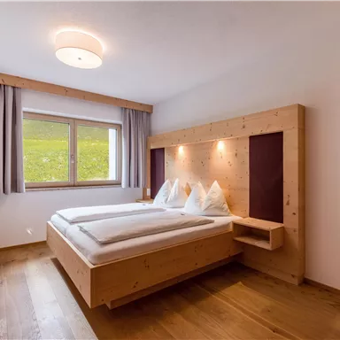 Ein modernes Schlafzimmer mit einem Doppelbett aus Holz. Große Fenster lassen viel Tageslicht herein und ermöglichen einen Blick ins Grüne.