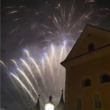 A beautiful fireworks display lights up the night sky behind a historical building. The bulb of a lantern shines in the foreground.