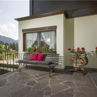 Ein einladender Balkon mit einer Sitzbank und bunten Blumen. Im Hintergrund sind sanfte Hügel und ein klarer Himmel zu sehen.