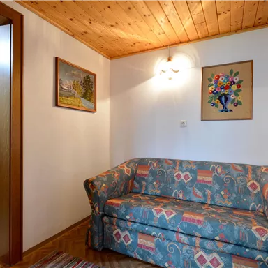 Ein gemütliches Wohnzimmer mit einem bunten Sofa und Holzboden. An der Wand hängen zwei Bilder, und im Hintergrund ist ein Badezimmer sichtbar.