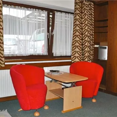 Ein gemütlicher Raum mit zwei roten Stühlen und einem kleinen Tisch. Die Fenster haben helle Vorhänge und bieten viel natürliches Licht.