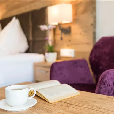 Ein gemütlicher Raum mit einem Holzregal und einem violetten Stuhl. Auf einem Tisch stehen eine Tasse Kaffee und ein geöffnetes Buch.