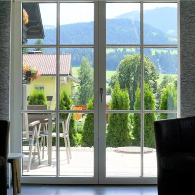 A view through a window of a green landscape with mountains in the background. In front of the window are two dark armchairs.