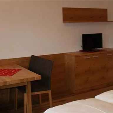 Ein gemütliches Zimmer mit einem Tisch, Stühlen und einem Fernseher. Die Wände sind hell, und es gibt Holzverkleidungen für eine warme Atmosphäre.