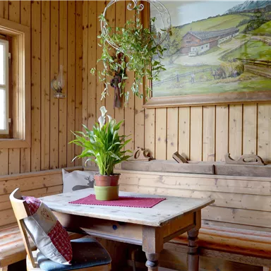 A cozy dining area with a wooden bench and a table. A landscape painting hangs on the wall, and there are plants for decoration.