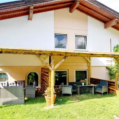 A cozy house with a covered terrace and a garden furniture set. The lawn is green and there are some plants in pots.