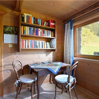 Ein gemütlicher Raum mit Holzwänden und einem kleinen Tisch. Eine Bücherregal und ein Fenster bieten einen Blick ins Grüne.