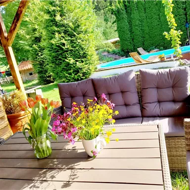 A cozy garden area with a table and comfortable seating cushions. In the background, a pool and lush green plants can be seen.