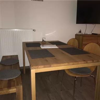 A modern dining area with a wooden tray and simple chairs. A television hangs on the wall, and there is a bench next to the table.