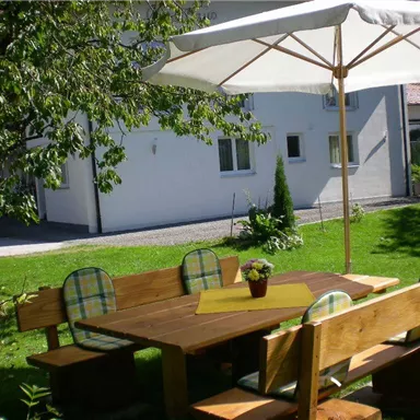 Ein gemütlicher Gartentisch aus Holz mit Bänken und einem Sonnenschirm. Umgeben von Grünflächen und Pflanzen in einem ruhigen Garten.