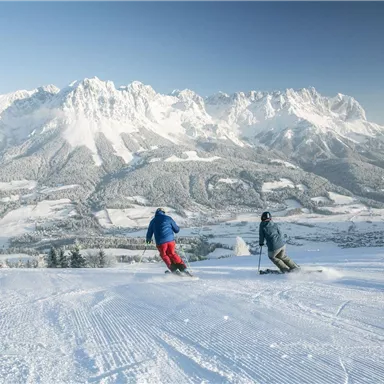 Two skiers are skiing down the slope, surrounded by snow-covered mountains. The clear sky and the snowy landscape create a wintry atmosphere.